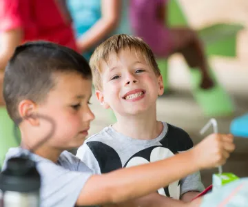 Cute boy looking at camera smiling while at lunch - friend is focused on his juice box