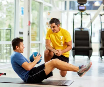 Male staff telling a male personal training client how to use the medicine ball whiling sitting on a yoga mat